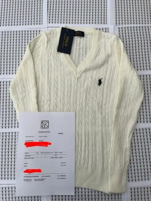 Vit kabelstickad tröja Polo Ralph Lauren - Kabelstickad vit tröja från Polo Ralph Lauren med V-ringning och klassisk broderad logga på bröstet. Tröjan har långa ärmar, ribbade muddar och är tillverkad i mjukt material som passar perfekt till lager på lager.