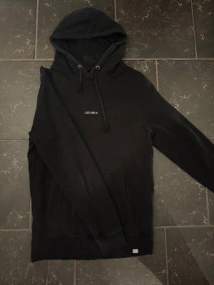 Svart hoodie från Les Deux - Snygg svart hoodie från Les Deux med minimalistisk vit logga på bröstet. Klassisk modell med huva och dragsnören samt magficka framtill. Tillverkad i mjukt material som känns skönt mot huden. Perfekt för en avslappnad och stilren look.