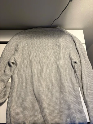 Ljusgrå stickad tröja från Gant - Snygg ljusgrå stickad tröja från Gant med diskret logga på bröstet. Tröjan har rund halsringning, ribbade muddar och är tillverkad i ett mjukt, strukturerat material. Perfekt för lager på lager och enkel att matcha.