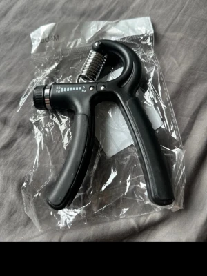 Svart handtränare  - Säljer en svart handtränare med justerbart motstånd och metallfjäder. Perfekt för att träna greppstyrka och underarmar. Ergonomiskt handtag i plast som är lätt att hålla i. Levereras i genomskinlig plastförpackning. 5-60kg går att justera.