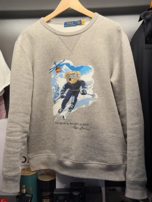 Grå sweatshirt Polo Bear Ralph Lauren - Grå sweatshirt från Ralph Lauren med Polo Bear-tryck där björnen åker skidor. Rund halsringning och ribbade muddar vid ärmslut och nederkant. Tröjan är helt ny! Finns bevis att den är äkta också. Nypris (3000)