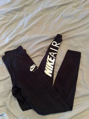 Svarta Nike Air leggings - Snygga svarta leggings från Nike Air med stor vit logga längs ena benet och mindre logga vid höften. Tight passform och stretchigt material som sitter skönt. Perfekta till träning eller chill. Klassisk sportig look med hög midja.