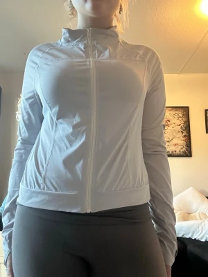 Vit sportjacka med dragkedja - Snygg vit sportjacka med hel dragkedja framtill och hög krage. Jackan har långa ärmar med tumhål och är tillverkad i ett stretchigt, tunt material som passar perfekt till träning eller löpning.