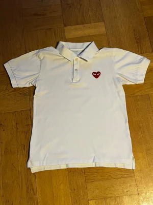 Vit pikétröja Comme des Garçons - Vit pikétröja från Comme des Garçons med klassisk krage och knäppning framtill. På bröstet sitter det ikoniska röda hjärta-märket. Tröjan har korta ärmar och är gjord i mjuk bomull. Säljer pga att den inte längre passar mig. 