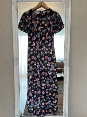 Lång blommig sammetsklänning med puffärm - Långklänning med svart botten och färgglatt blommönster i rosa, lila och grönt. Klänningen har korta puffärmar och rund halsringning. Perfekt för dig som vill ha en romantisk och trendig look.