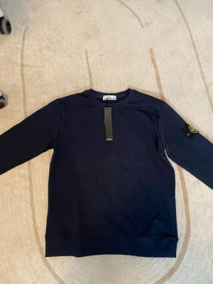 Mörkblå stickad tröja Stone Island - Mörkblå stickad tröja från Stone Island med klassisk rund halsringning och ribbade muddar. Tröjan har den ikoniska logotypen på vänster ärm och är tillverkad i mjukt material som passar perfekt till en avslappnad stil.