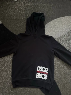 Dsquared2 Hoodie - Snygg svart hoodie från Dsquared2 med stor huva och tryck i vitt och rött på nedre delen av framsidan. Tillverkad i mjukt bomullsmaterial som känns skönt mot huden. Perfekt för dig som gillar streetwear och vill ha något stilrent men ändå med attityd. Knappt använd. STLRK 14Y