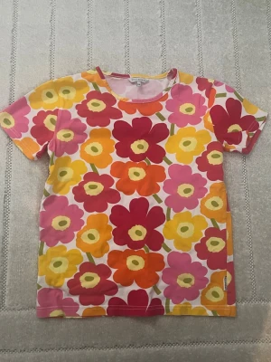 Blommig t-shirt från Marimekko - Färgglad t-shirt från Marimekko med stort blommigt mönster i rosa, gul, orange och röd. Klassisk rund halsringning och korta ärmar. Perfekt för dig som gillar statement-plagg och vill sticka ut med en ikonisk design.