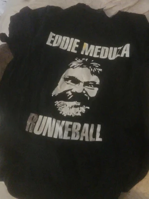 Svart Eddie Meduza t-shirt - Säljer en svart t-shirt med tryck av Eddie Meduza och texten 'RUNKEBALL' i vitt. T-shirten har klassisk passform och rund hals. Perfekt för dig som gillar ikoniska bandmotiv och vill sticka ut med en unik tröja.