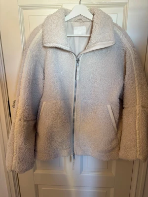 Vit teddyjacka med dragkedja - Mysig vit teddyjacka med dragkedja framtill och två stora fickor. Jackan har en hög krage och breda ärmar, samt detaljer längs ärmarna. Perfekt för att hålla värmen och ge en trendig look. Materialet är fluffigt och mjukt, typiskt för teddy.