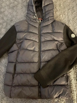 Svart pufferjacka från Moncler - Säljer en svart pufferjacka från Moncler med quiltad kropp och stickade ärmar. Jackan har huva med dragsko, ribbade muddar och dragkedja framtill. Moncler-logga på ena ärmen. Perfekt för kalla vinterdagar och riktigt snygg streetstil.