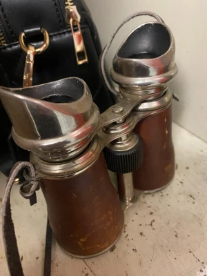 Vintage kikare med läderdetaljer - Säljer en klassisk vintage kikare med bruna läderdetaljer och metall i silverfärg. Kikaren har justerbart hjul i mitten och en rem för att hänga runt halsen. Perfekt för dig som gillar retroprylar eller samlar på gamla optiska instrument.