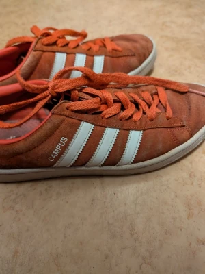 Adidas Campus orange sneakers - Säljer ett par Adidas Campus sneakers i orange mocka med klassiska vita ränder på sidorna och matchande orange skosnören. Låg modell med vit sula och Campus-tryck på sidan. Perfekta för dig som gillar retrostil och vill sticka ut med färg.