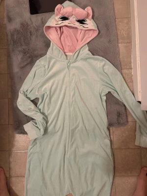 Mintgrön mjuk onesie med katt-hood - Supermysig mintgrön onesie med dragkedja och huva som har ett gulligt kattansikte, öron och rosett i ljusrosa. Insidan av huvan är ljuslila. Perfekt för chill hemma eller sleepover. Materialet är mjukt och känns som fleece.