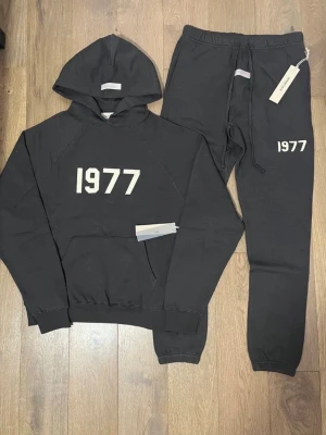 Svart loungewear-set 1977 Essentials - Snyggt svart loungewear-set från Essentials med trycket '1977' i vitt på både hoodie och byxor. Hoodien har känguruficka och dragsko i huvan, medan byxorna har resår i midjan och muddar vid bensluten. Perfekt för chill dagar hemma/skolan och ba ute!