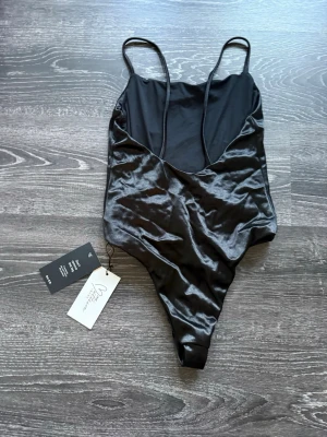 Svart satinbody från NA-KD - Snygg svart body från NA-KD i glansigt satinmaterial. Modellen har tunna axelband och djup urringning bak. Perfekt att styla med jeans eller kjol för en trendig look. Lappen sitter kvar.