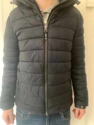 Mörkblå pufferjacka från superdry - Snygg mörkblå pufferjacka med huva och dragkedja framtill. Jackan har två sidofickor med dragkedja och quiltad design som ger en modern look. Perfekt och enkel att matcha med olika outfits. Några små defekter, se på bild 4,5,6 och 7. Sitter bra på mig och jag är 180.