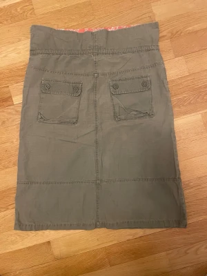 Olivgrön långkjol med fickor y2k vintage - Säljer en olivgrön långkjol med två stora fickor bak som har lock och knappar. Kjolen har en rak passform och är sydd i ett mjukt bomullstyg. Perfekt för dig som gillar en avslappnad och enkel stil.