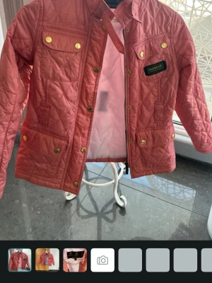 Quiltad rosa jacka från Barbour - Säljer en quiltad rosa jacka från Barbour med guldfärgade knappar och dragkedja. Jackan har flera fickor framtill och ett Barbour-märke på bröstet. Perfekt för dig som vill ha en snygg och bekväm jacka till vardags.