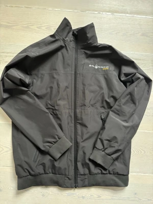 Svart vindjacka från Sail Racing - Svart vindjacka från Sail Racing med dragkedja framtill och två sidofickor med dragkedja. Jackan har hög krage, ribbade muddar och diskret logga på bröstet. Materialet är tunt och vindtätt, perfekt för blåsiga dagar.