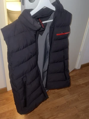 Svart dunväst från Prada - Snygg svart dunväst från Prada med quiltad design och hög krage. Västen har en röd detalj med Prada-logga på bröstet och dragkedjeförsedda fickor. Perfekt för lager-på-lager och streetwear-stil. Dragkedjan går tyvärr inte att stänga, litet hål precis under Prada märket som också går att laga därav är priset satt ganska lågt!