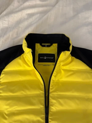 Gul och svart dunväst från Sail Racing - Snygg dunväst från sail Racing i en cool gul och svart färgkombination. Västen har hög krage, dragkedja framtill och quiltad design. Perfekt för lager-på-lager och ger en sportig vibe till din outfit.