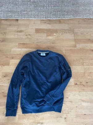 Mörkblå sweatshirt från C.P. Company - Snygg mörkblå sweatshirt från C.P. Company med rund halsringning och ikonisk linsdetalj på ärmen. Tröjan har en avslappnad passform och ribbade muddar vid ärmslut och nederkant. Perfekt för en casual streetwear-look.