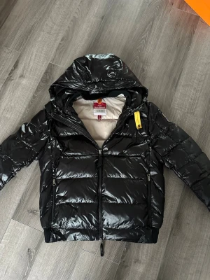 Parajumpers sheen svart - Snygg svart dunjacka från Parajumpers sheen med glansig finish och huva. Jackan har dragkedja framtill, två sidofickor med dragkedja och en gul detalj på dragkedjan. Perfekt för kalla vinterdagar och riktigt trendig look.
