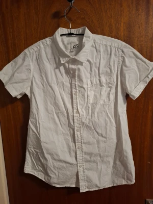 Vit kortärmad skjorta från Slim 63 - Klassisk vit skjorta från Slim 63 med korta ärmar och bröstficka. Skjortan har knappar framtill och en enkel, rak passform. Perfekt till sommaren och lätt att matcha med olika outfits.