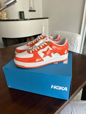 BAPE STA orange och vita sneakers - HELT OANVÄNDA! Dom var försmå för mig.. Säljer ett par BAPE STA sneakers i orange och vitt med glansig finish. Skorna har den ikoniska stjärnan på sidan, vita snören och perforerad tå. Klassisk low-top siluett och gummisula. Perfekta för dig som vill sticka ut med streetwear-stil.