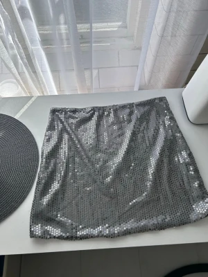 Glittrig silver paljettkjol - Säljer en kort, glittrig silverkjol täckt med paljetter. Kjolen har en rak passform och är fodrad på insidan för extra komfort. Perfekt för dig som vill sticka ut med en riktigt snygg partylook!