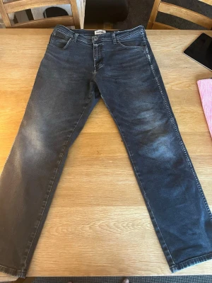 Mörkblå jeans från Wrangler - Snygga mörkblå jeans från Wrangler med klassisk femficksmodell och raka ben. Jeansen har en lätt tvättad look med slitningar på framsidan och tydliga sömmar på bakfickorna. Perfekta för en avslappnad och trendig stil.