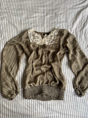 Olivgrön blus med spets från Vero Moda - Snygg olivgrön blus från Vero Moda i lätt material. Blusen har långärmade puffärmar, bred smock i midjan och vacker spetsdetalj med knappar vid halsen. Perfekt för dig som gillar en avslappnad men chic stil.