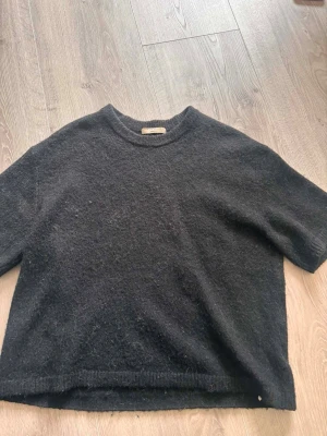 Mörkgrå stickad t-shirt - Mysig mörkgrå stickad t-shirt med korta ärmar och rund hals. Tröjan har en avslappnad passform och är tillverkad i ett mjukt, luddigt material som ger en skön känsla. Perfekt för lager på lager och enkel att styla till olika outfits.