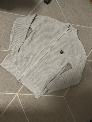 Grå stickad ziptröja från Prada - Snygg grå stickad ziptröja från Prada med hög krage och ikonisk trekantslogga på bröstet. Tröjan har grovstickad struktur, långa ärmar och dragkedja framtill. Perfekt för dig som gillar exklusiva och stilrena plagg.