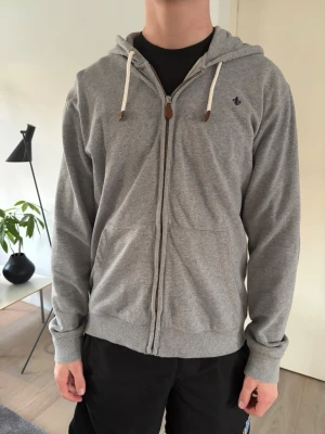 Grå Morris Zip Hoodie - Säljer en grå hoodie från Morris med dragkedja framtill och vita dragsnören i huvan. Hoodien har två fickor fram och en liten broderad logga på bröstet. Perfekt för chill dagar och enkel att matcha med det mesta.