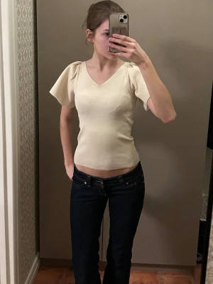 Krämvit topp med puffärmar - Säljer en krämvit topp med v-ringning och korta puffärmar. Toppen är figurnära och har en mjuk, stretchig känsla. Perfekt för dig som gillar en enkel men trendig stil. Använd fåtal gånger