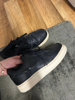 Nike Air Force 1 svart/vit sneakers - Säljer ett par klassiska Nike Air Force 1 sneakers i svart läder med vita sulor. Skorna har svarta skosnören och en diskret regnbågsskimrande detalj på sidan. Perfekta för dig som gillar stilrena och tidlösa sneakers.