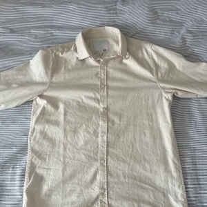 Beige skjorta från 157, storlek M - Stilren beige skjorta från 157 i storlek M. Skjortan har klassisk krage, knäppning framtill med vita knappar och är tillverkad i ett lätt bomullstyg. Perfekt för en clean och enkel look.