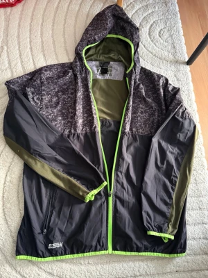 Svart och grön vindjacka  - Snygg vindjacka  i svart och olivgrönt med digitalt camomönster upptill. Jackan har huva, hel dragkedja med limegröna detaljer och två fickor med dragkedja. Tillverkad i lätt och vindtätt material, perfekt för blåsiga dagar.