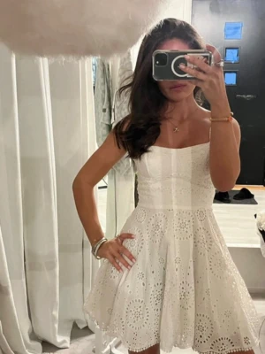 Vit broderad kortklänning med spets - Söt vit kortklänning med broderade spetsdetaljer och axelband. Klänningen har en utsvängd kjol och markerad midja, perfekt till midsommar eller studenten