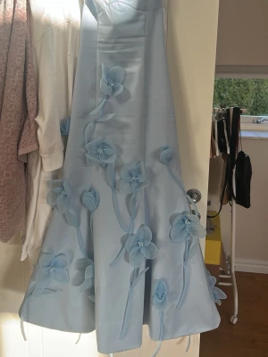 Ljusblå axelbandslös klänning med blommor - Superfin ljusblå axelbandslös klänning med 3D-blommor längs hela framsidan. Klänningen har en elegant, utsvängd form och är gjord i ett glansigt material som ger en lyxig känsla. Perfekt för speciella tillfällen och student.