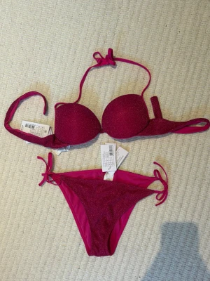 Glittrig rosa bikini från Calzedonia - Snygg rosa bikini från Calzedonia med glittrigt tyg. Överdelen har vadderade kupor och knytband i nacken (alternativt rakt bak som en vanlig bh, beror på hur man vill ha den), medan underdelen har knyt i sidorna för justerbar passform. Perfekt för strandhäng eller poolparty!