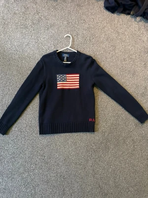 Mörkblå stickad tröja Polo Ralph Lauren - Mörkblå stickad tröja från Polo Ralph Lauren med amerikansk flagga broderad på bröstet och RL-broderi i rött vid nederkanten. Tröjan har rund halsringning, långa ärmar och ribbstickade muddar. Perfekt för dig som gillar klassisk stil.