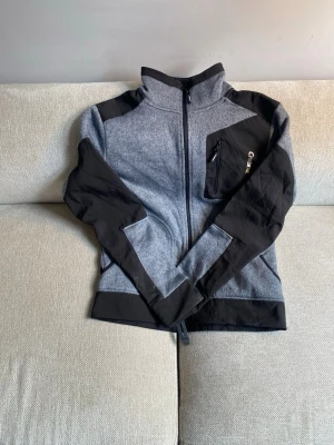 Grå och svart fleecejacka Cratex - Sportig grå och svart fleecejacka från Cratex med hel dragkedja framtill, hög krage och tryck i rött på ryggen. Jackan har svarta detaljer på axlar och ärmar samt en bröstficka med dragkedja. Perfekt för lager-på-lager under kyliga dagar.