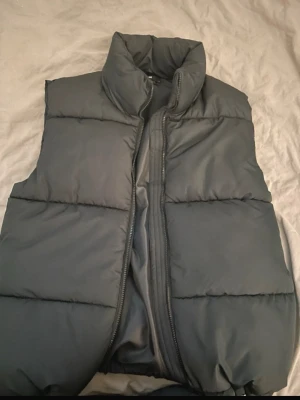 Svart dunväst med hög krage - Stilren svart dunväst med hög krage och dragkedja framtill. Västen har breda, vadderade paneler och en enkel, minimalistisk design som passar till många olika outfits. Perfekt för lager-på-lager och håller dig varm under kyliga dagar.