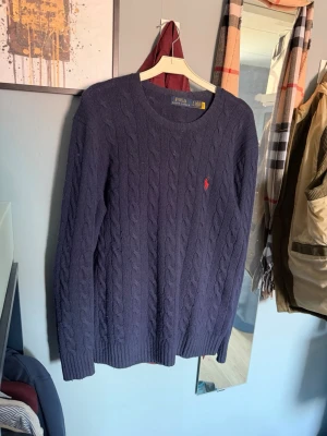 Mörkblå kabelstickad tröja Polo Ralph Lauren - Snygg mörkblå kabelstickad tröja från Polo Ralph Lauren med rund halsringning och den klassiska röda logotypen broderad på bröstet. Tröjan har långa ärmar och ribbade muddar vid ärmslut och nederkant. Perfekt för en stilren look.