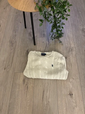 Vit kabelstickad tröja Polo Ralph Lauren - Kabelstickad vit tröja från Polo Ralph Lauren med rund halsringning och den klassiska blå logotypen broderad på bröstet. Tröjan har långa ärmar och ribbade muddar vid ärmslut och nederkant. Perfekt för en clean och stilren look.