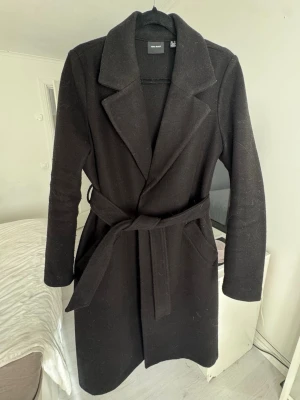Svart lång kappa från Vero Moda - Stilren svart kappa från Vero Moda med klassisk krage och knytskärp i midjan. Kappan har raka ärmar och två sidofickor. Materialet är tjockt och känns som ullblandning, perfekt för kyliga dagar. Enkel och tidlös design som passar till många olika outfits.