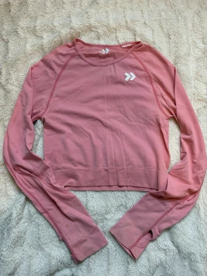 Rosa långärmad träningströja  - Säljer en långärmad topp i rosa med rund hals och diskret logga. Stretchigt material, passar både träning och vardag. Står ingen storlek men skulle säga XS/XXS. En liten fläck på sista bilden kan troligen försvinna i tvätten 